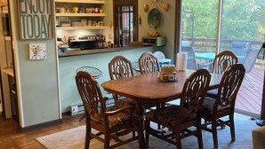 Uma sala de jantar com uma mesa redonda e seis cadeiras, uma cozinha com diversos eletrodomésticos e decoração, e uma grande janela com vista para o exterior.
