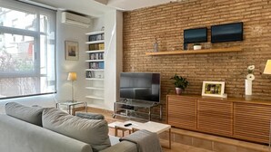 Ein modernes Wohnzimmer mit einem grauen Sofa, einem Fernseher auf einem Schrank, einem Bücherregal und einer Ziegelwand.