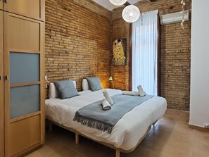 Un dormitorio con pared de ladrillo, cama con ropa de cama blanca y almohadas grises, armario de madera, un cuadro y un equipo de aire acondicionado.