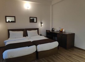 Uma cama de casal com cabeceira de madeira, mesa de cabeceira e escrivaninha com cadeira. Há duas luminárias fixadas na parede e um espelho sobre a cama.