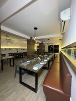 A modern restaurant with long communal tables, wooden pillars, and hanging pendant lights.