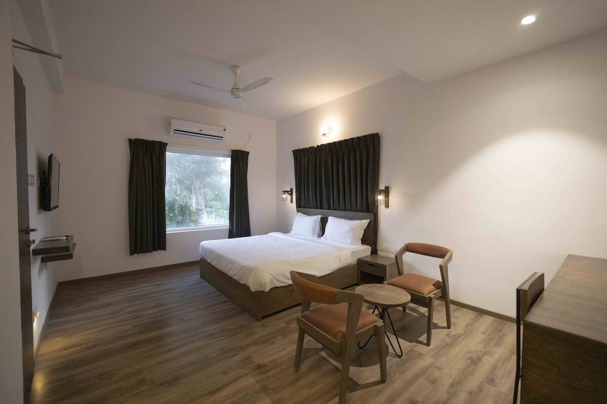 A hotel room with a bed, a desk, two chairs, a ceiling fan, an air conditioning unit, and a window with curtains.