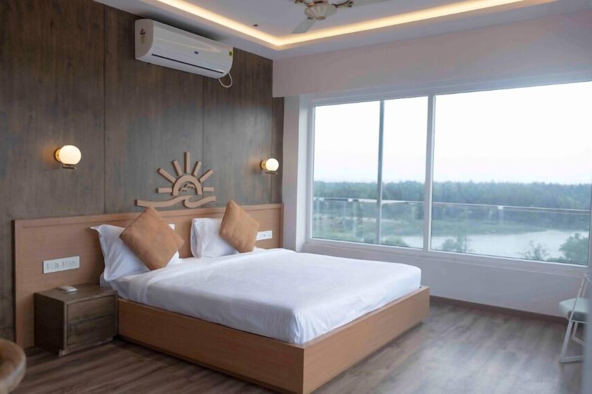 A modern hotel room with a large bed, wooden headboard, two wall-mounted lights, and a view of a bridge through a large window.