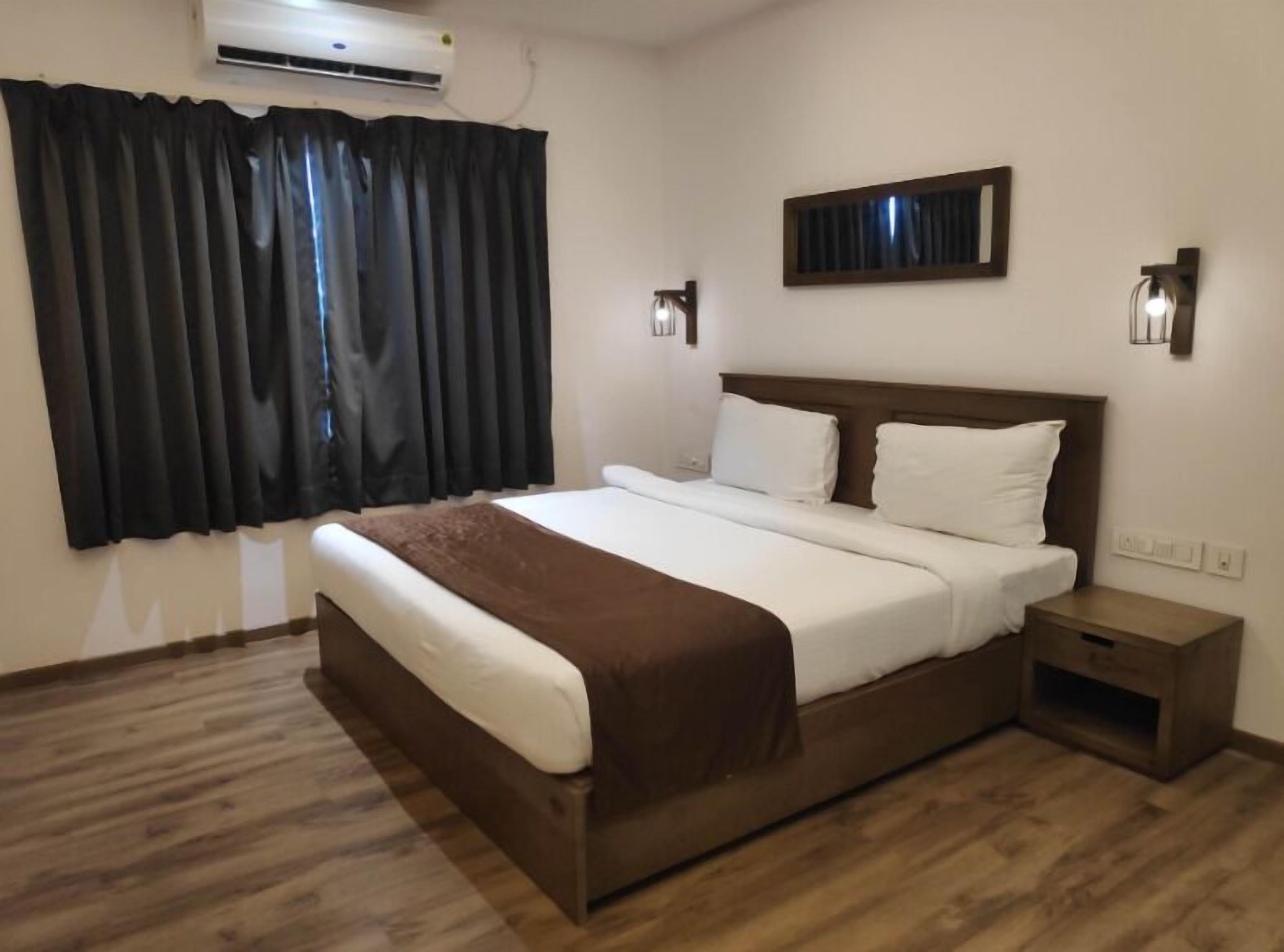 A hotel room with a large bed, a bedside table, a nightstand, a wall-mounted air conditioner, and a window with dark curtains.