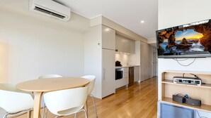 A modern kitchen with white cabinets, a built-in oven, and a dishwasher. There is a dining area with a round table and white chairs. A flat-screen TV is mounted on the wall above a wooden shelf. - The Sand Resort Kingscliff - One Bed (8308) (Tweed)