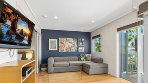 A modern living room with a grey sectional sofa, a flat-screen TV, wooden flooring, and a balcony with a view of palm trees. - The Sand Resort Kingscliff - One Bed (8308) (Tweed)