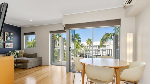 A modern living room with a sofa, a dining table with chairs, and a sliding glass door leading to a balcony with a view of palm trees and buildings. - The Sand Resort Kingscliff - One Bed (8308) (Tweed)