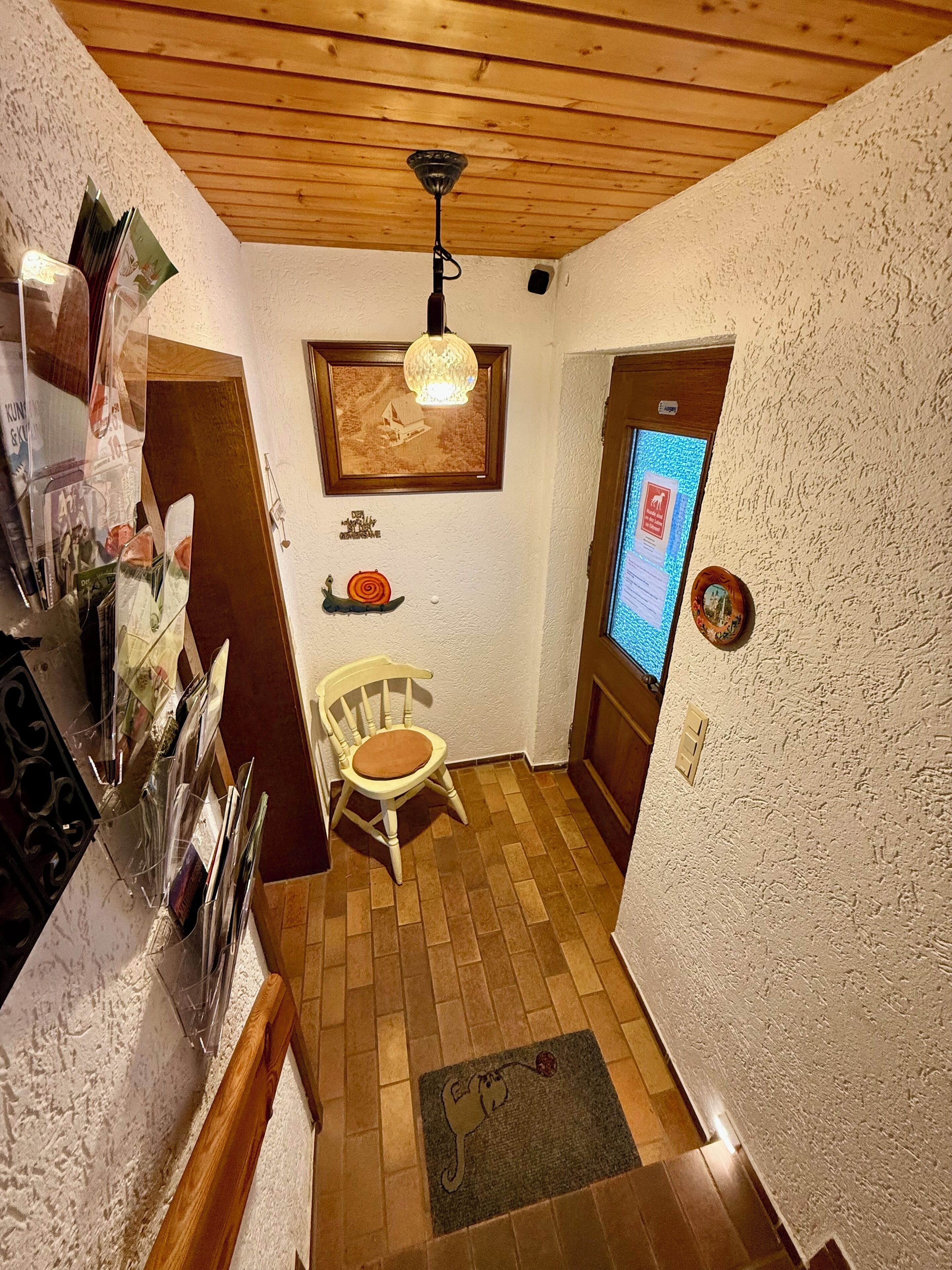 Ein kleiner, gemütlicher Raum mit Holzbalkendecke, einem gerahmten Bild, einem Tisch mit Stuhl und einem digitalen Display an der Wand.