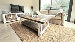 A living room with a wooden coffee table, a sofa, a TV on a white cabinet, and a large window with a view of trees.