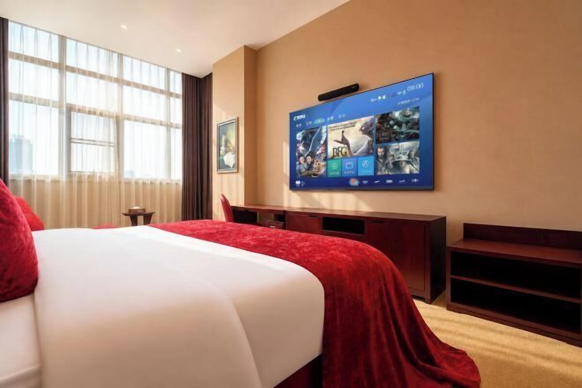 A hotel room with a large bed, a TV mounted on the wall, a desk, and a window with curtains.