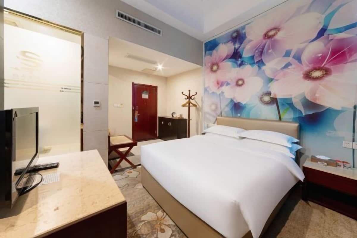 Ein Hotelzimmer mit einem Bett, einem Schreibtisch mit Computer, einem Kleiderhaken und einer Wand mit floralem Wandbild.