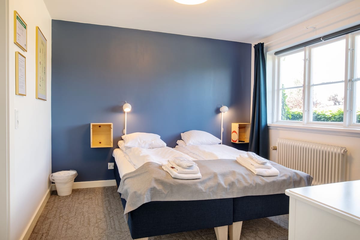 Hotellihuone, jossa on parivuode, kaksi yöpöytää, pieni puinen kaappi, patteri ja ikkuna, jossa on verhot.