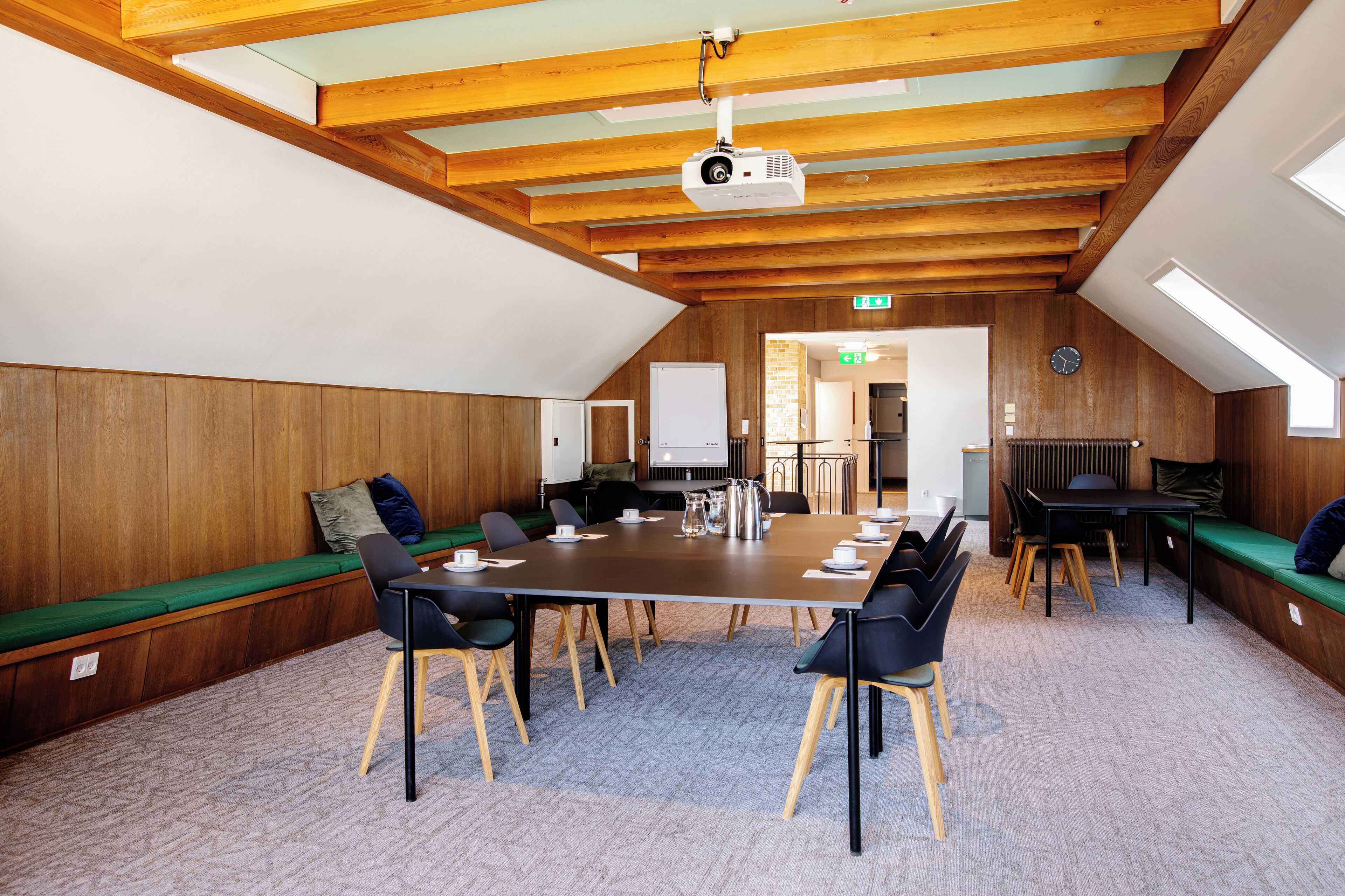 A conference room with a long table, chairs, a projector, and a view of the kitchen through an open door.