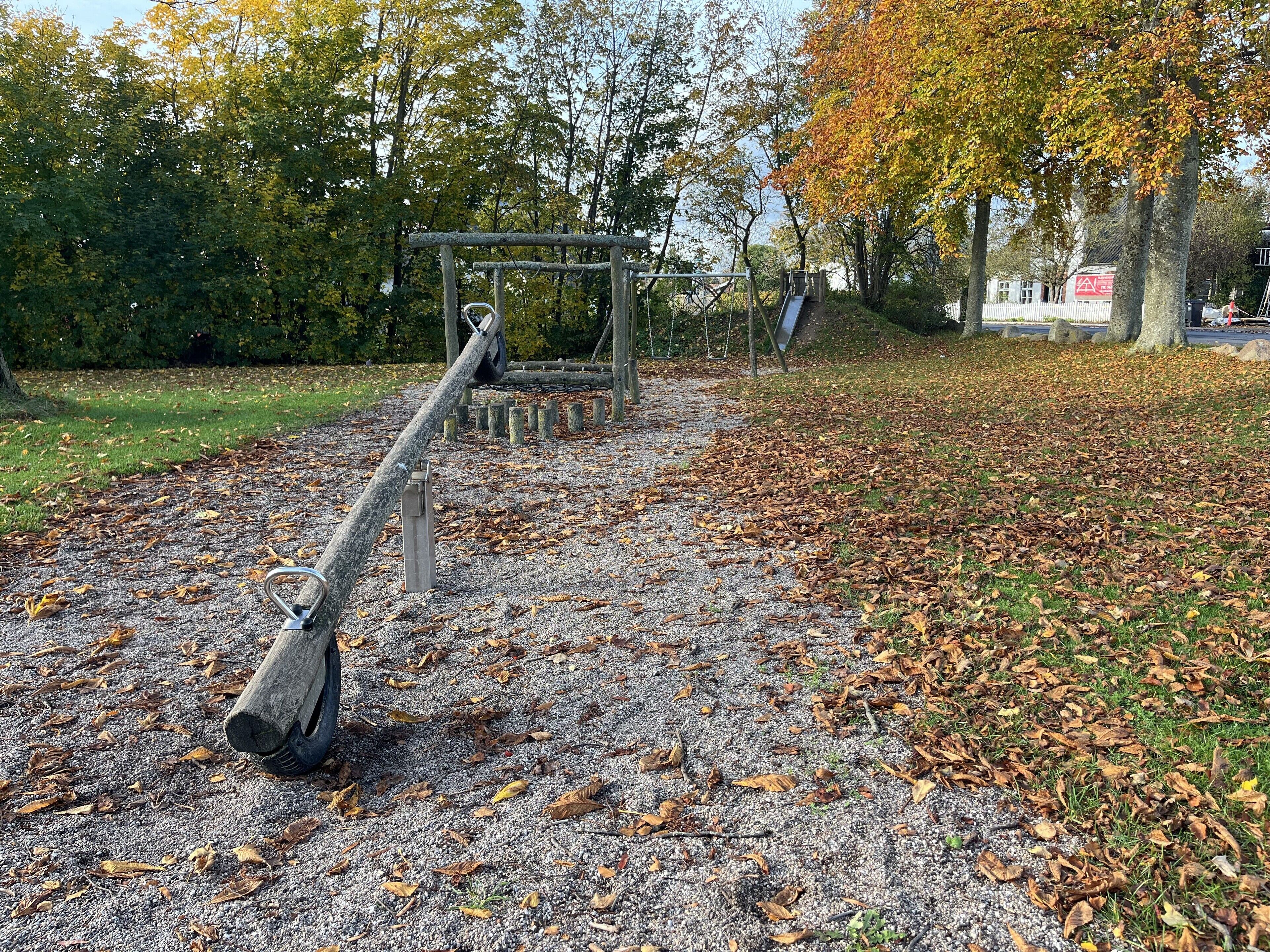 A playground with a seesaw and a slide, surrounded by trees and a gravel path.