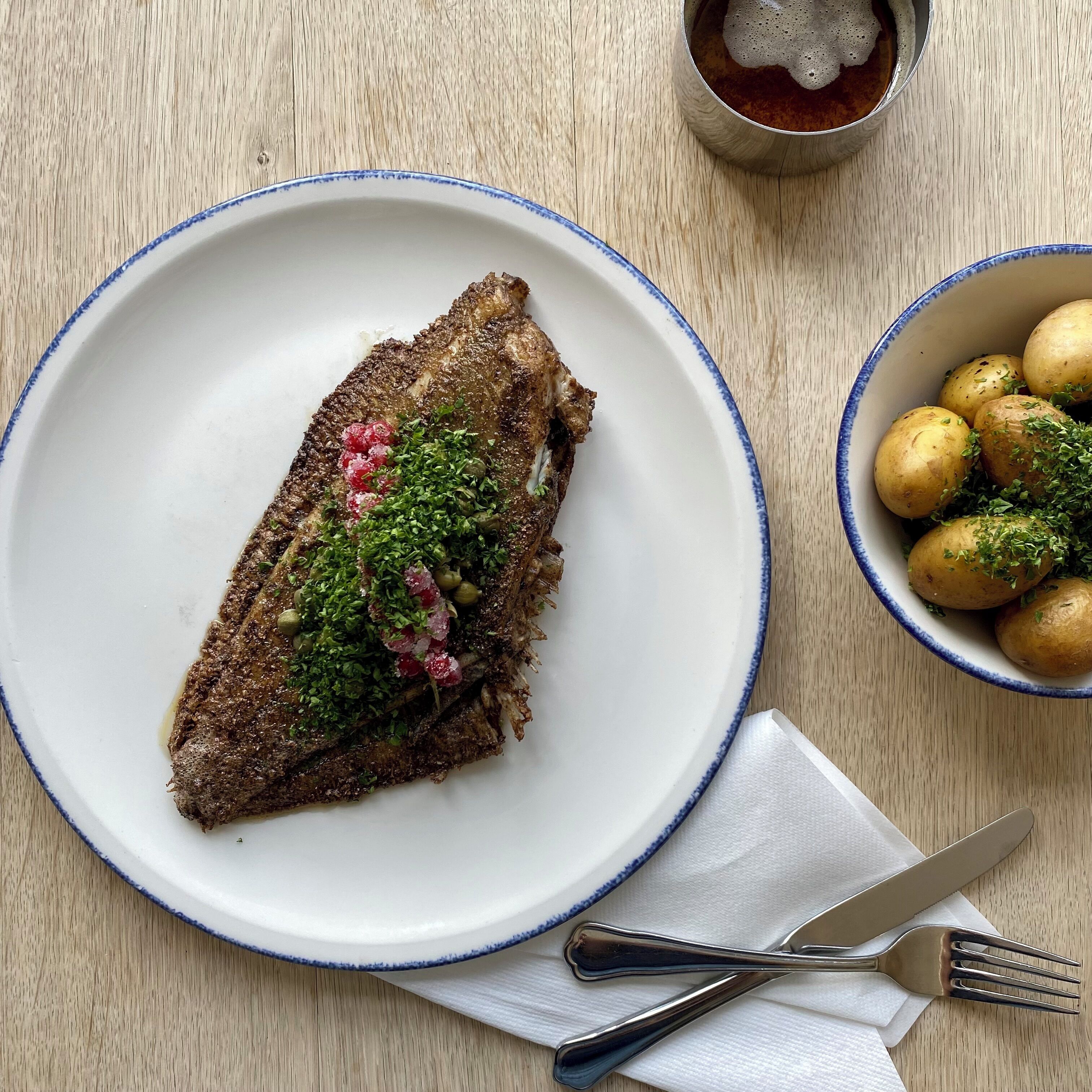 Une assiette avec un poisson, un bol de pommes de terre, et une fourchette et un couteau sur une table en bois.