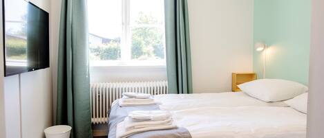 A hotel room with two beds, a TV, a radiator, a lamp, and a window with curtains.