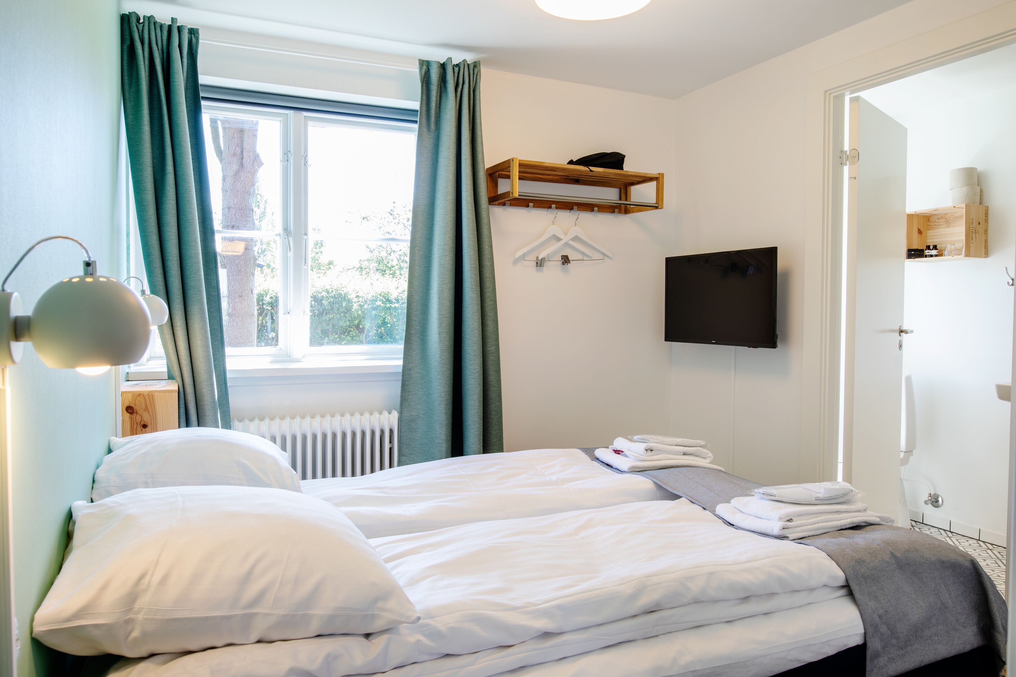 Una habitación de hotel con una cama, una mesita de noche con lámpara, un televisor montado en la pared y una ventana con cortinas.