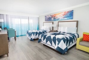 A hotel room with two beds, a desk, chairs, and a large window with sheer curtains.