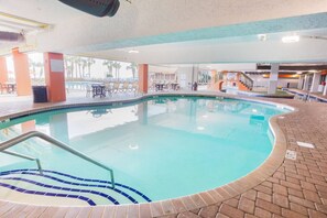 An indoor swimming pool with a tiled edge, a set of stairs leading into the pool, and a lounge area with tables and chairs.