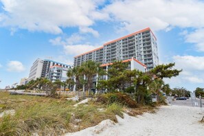 A beachfront hotel with multiple floors, a sandy beach in the foreground, and a clear sky.
