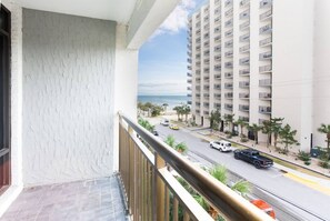 A balcony with a view of a multi-story building, a road with vehicles, and a beach in the distance.