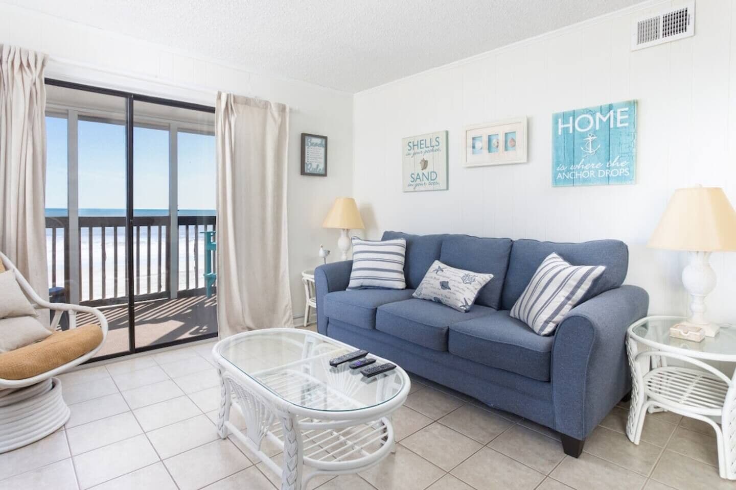 Una sala de estar con un sofá azul, una mesa de centro de vidrio, un sillón blanco y vista a la playa a través de una puerta corrediza de vidrio.