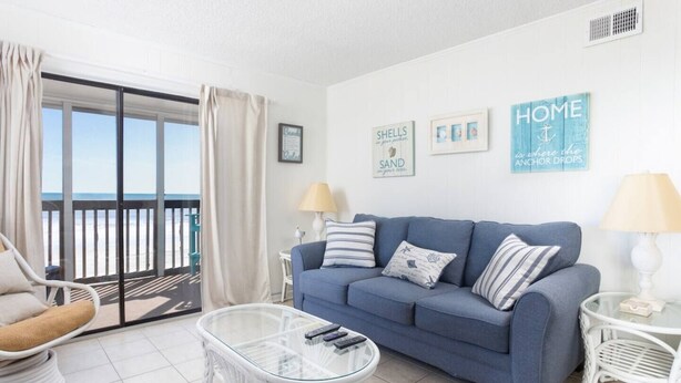 A living room with a blue sofa, a glass coffee table, a white armchair, and a view of the beach through a sliding glass door.