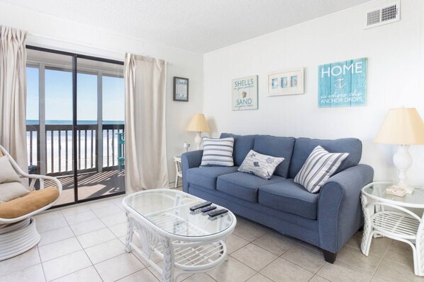 A living room with a blue sofa, a glass coffee table, a white armchair, and a view of the beach through a sliding glass door.