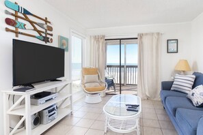 Een woonkamer met een televisie, witte meubels, een glazen tafel en uitzicht op het strand vanuit een balkon.