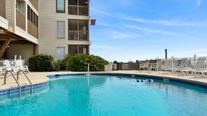 A swimming pool with a clear blue water surface, surrounded by a white fence, and a building with balconies and a red flag.