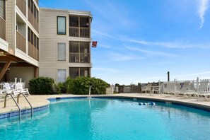 A swimming pool with a clear blue water surface, surrounded by a white fence, and a building with balconies and a red flag.