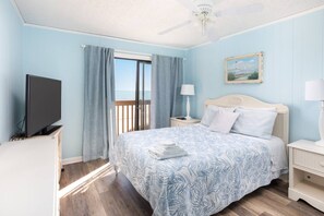 A bedroom with a bed, a television, a nightstand, a lamp, a painting, and a view of the outdoors.