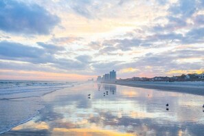 Een strand met een rustige zee, nat zand dat de hemel weerspiegelt en gebouwen in de verte.