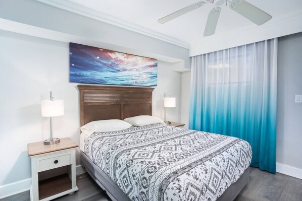 Une chambre d’hôtel avec un lit, des tables de chevet, un ventilateur de plafond, un tableau et une fenêtre avec des rideaux.