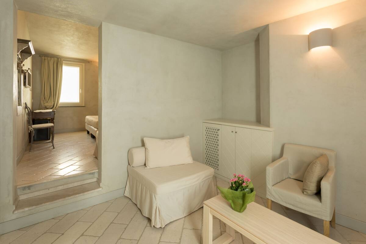 A small, well-lit room with a bed, a chair, a table, and a cabinet.