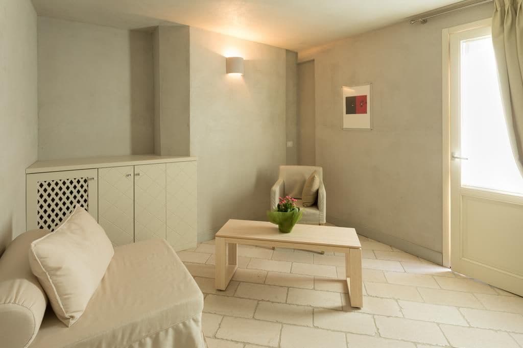 Habitación en tonos neutros con un sofá, una mesa de centro de madera, una silla, una lámpara de pared y una ventana con cortinas.