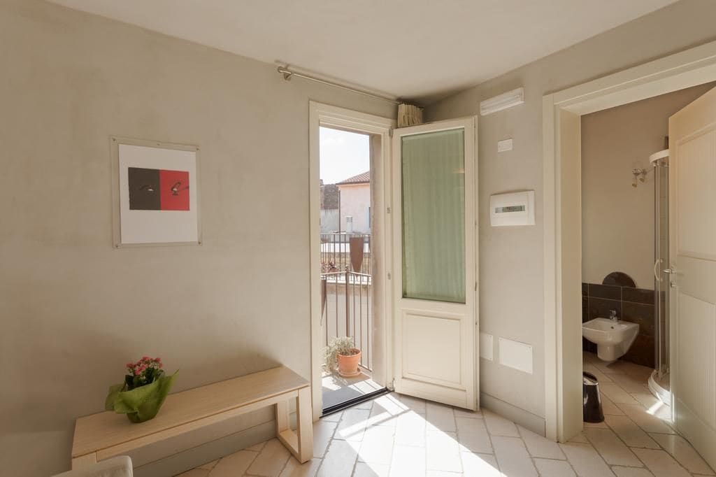 A room with a bench, a door leading to a balcony, a bathroom with a bidet, and a framed picture on the wall.