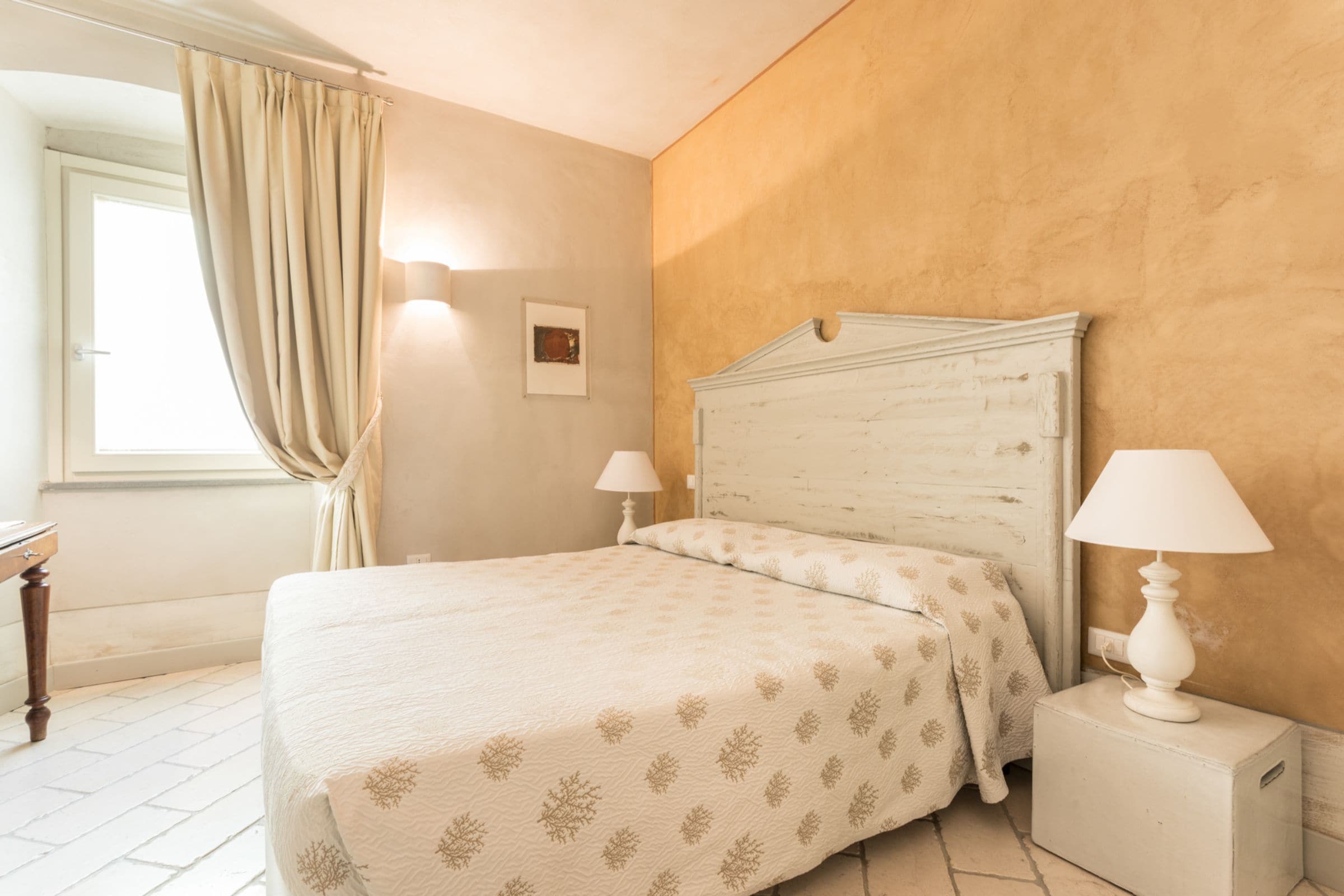 Un dormitorio con cama, dos lámparas de noche, una mesita de noche, una ventana con cortinas y un cuadro colgado en la pared.