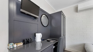 Une chambre d’hôtel moderne avec un bureau gris, un téléviseur à écran plat, une machine à café et un lit placé contre un mur en briques blanches.