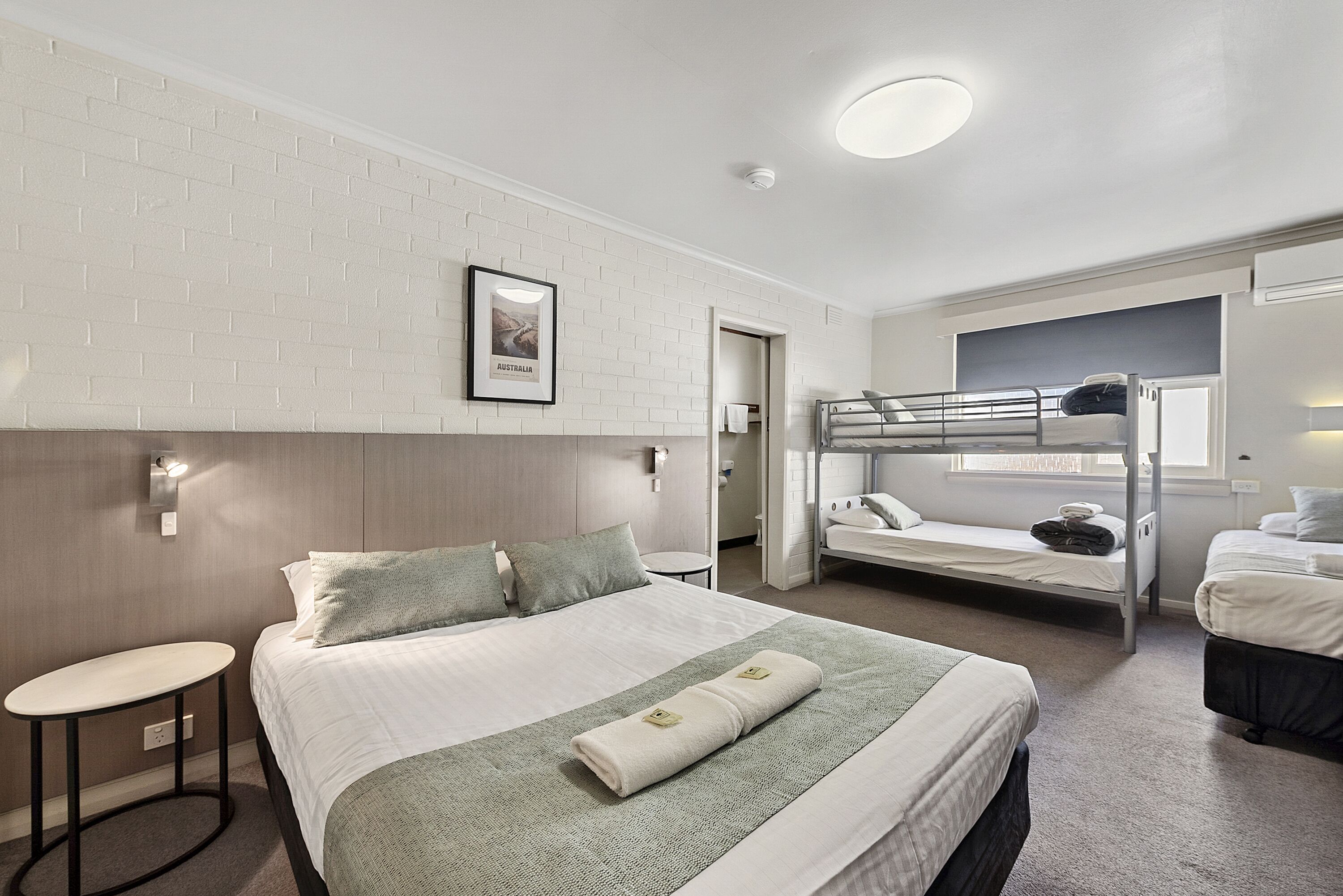 A hotel room with a bunk bed, a single bed, a small table, and a framed picture on the wall.