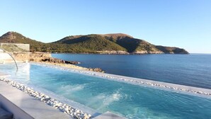 On aperçoit clairement une piscine bordée de rochers, une mer calme et, au loin, un paysage vallonné.
