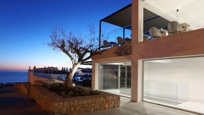 A modern building with a glass facade, a stone planter with a tree, and a view of the sea. - Mar Azul Hotel - Adults Only (Capdepera)