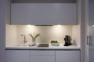 A modern kitchen with a white countertop, a built-in sink, a coffee machine, a bowl of green apples, a potted plant, and a white telephone.