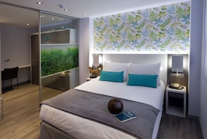 A modern hotel room with a large bed, a desk, and a view of greenery through a glass door.