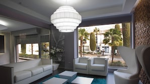 Een moderne woonkamer met een bank, twee fauteuils en een salontafel. Er is een groot raam met uitzicht op palmbomen en een gebouw.