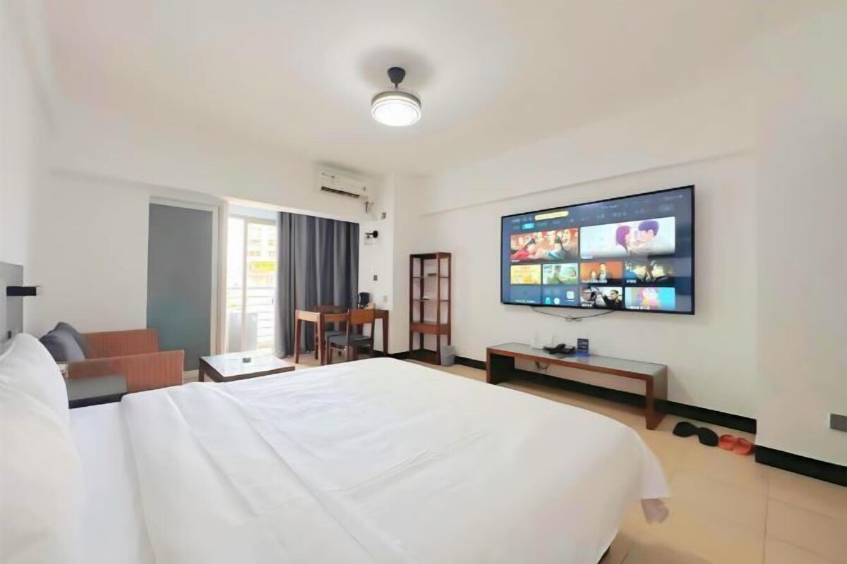A hotel room with a large bed, a desk, a chair, a TV mounted on the wall, and a window with curtains.