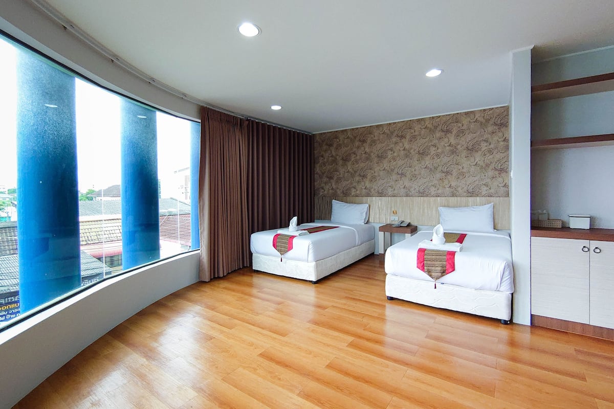 Habitación de hotel con dos camas, un ventanal con cortinas, suelo de madera y papel tapiz estampado en la pared.
