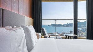 A hotel room with a large bed, a chair, a small table, and a view of the sea and cityscape through large windows. - Hotel The Club Yeonhwari (Busan)