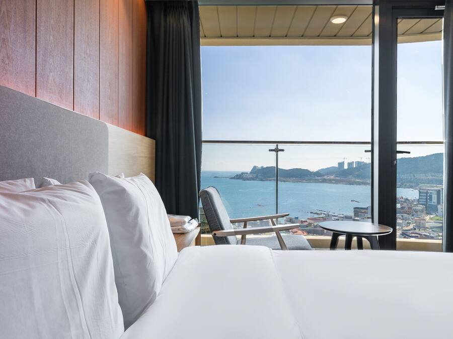 A hotel room with a large bed, a chair, a small table, and a view of the sea and cityscape through large windows.