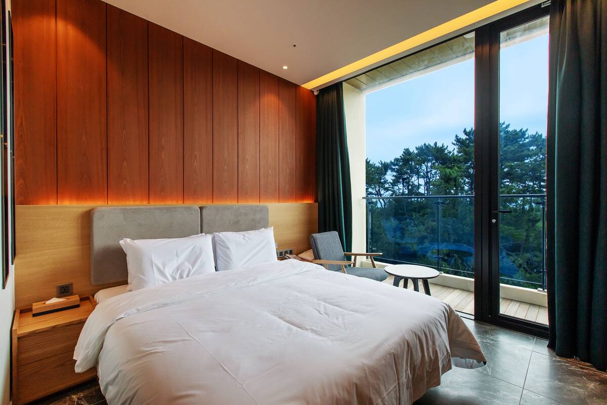 A modern hotel room with a large bed, a small table, and a balcony with a view of trees.